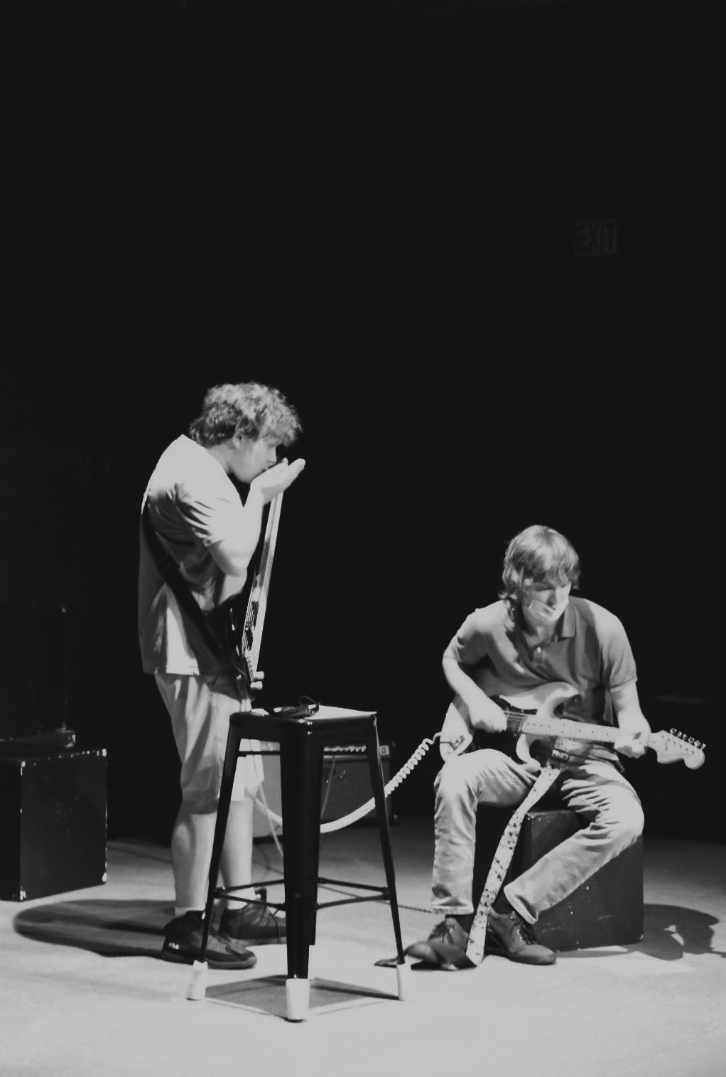 One student stands to the left playing a harmonica and holding a guitar; another student sits to the right playing a guitar.